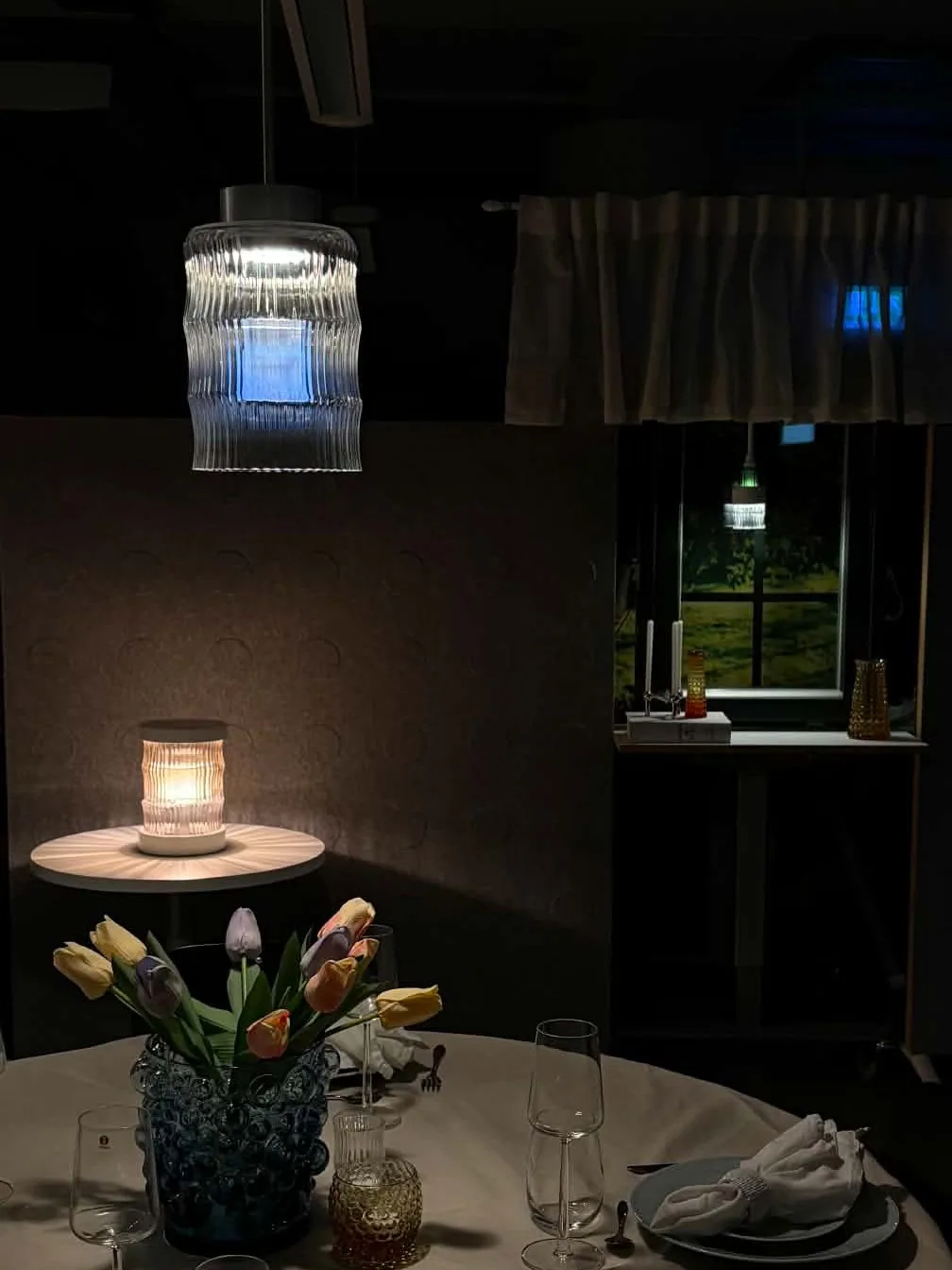 Celina lit over a laid dining table at the exhibition, with a table lamp and a window pendant behind it